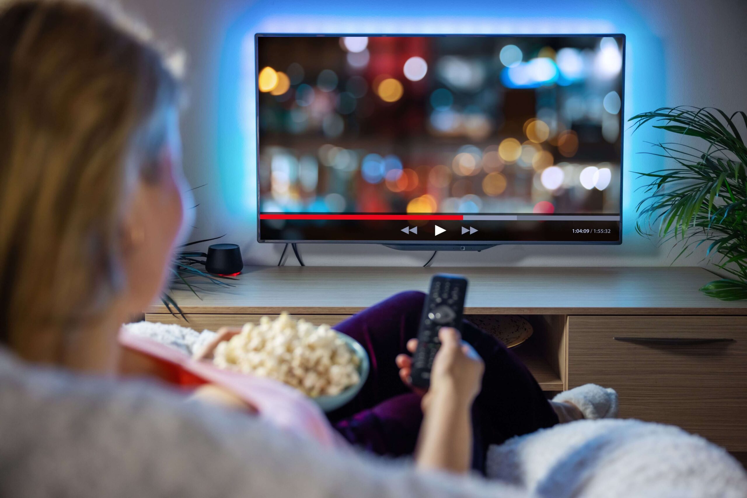 A person relaxes on a couch, holding a remote and popcorn while watching blurred content on a wall-mounted TV screen.