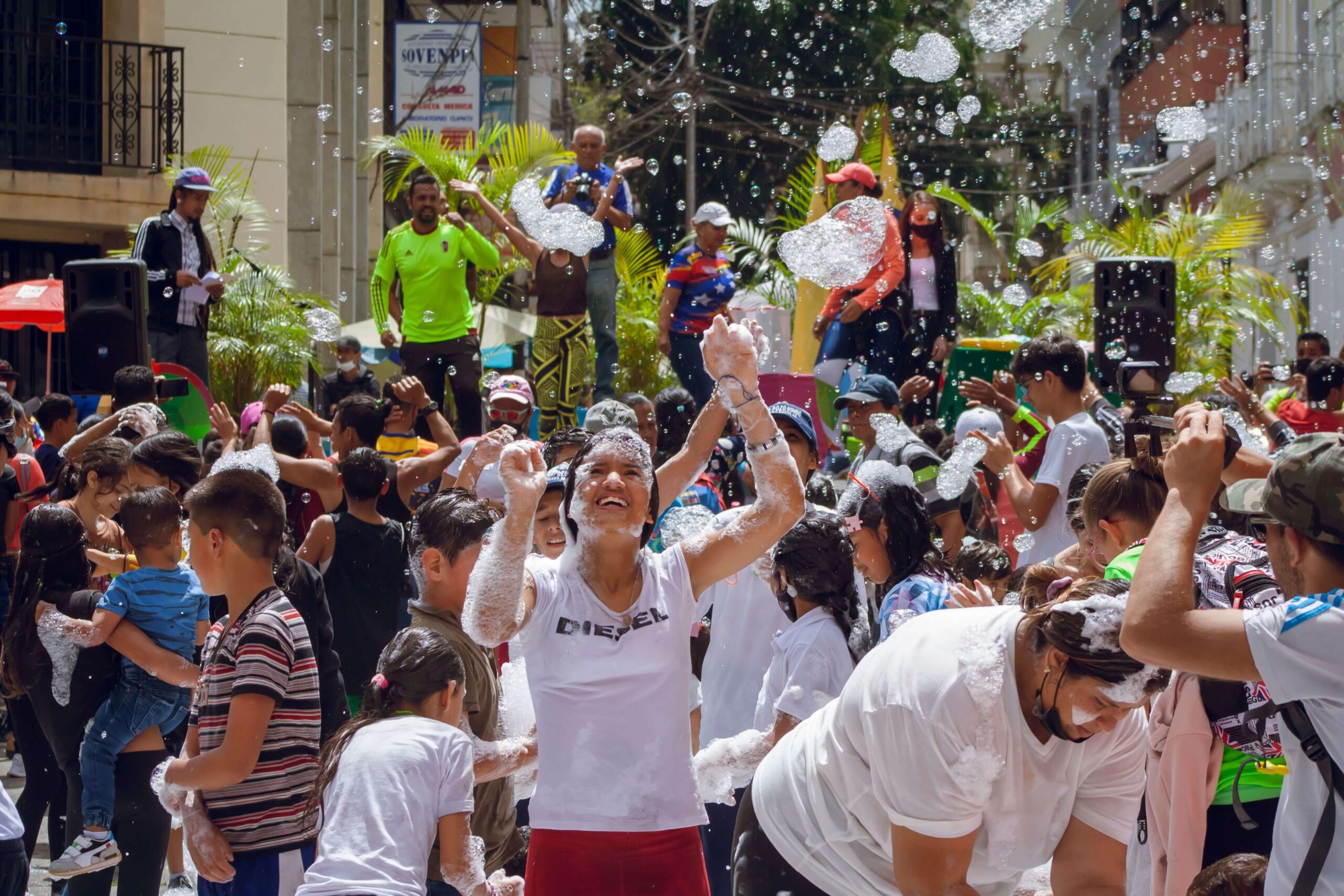 A lively foam party with joyful people, children, and bubbles in an urban setting. No recognizable landmarks or historical buildings visible.
