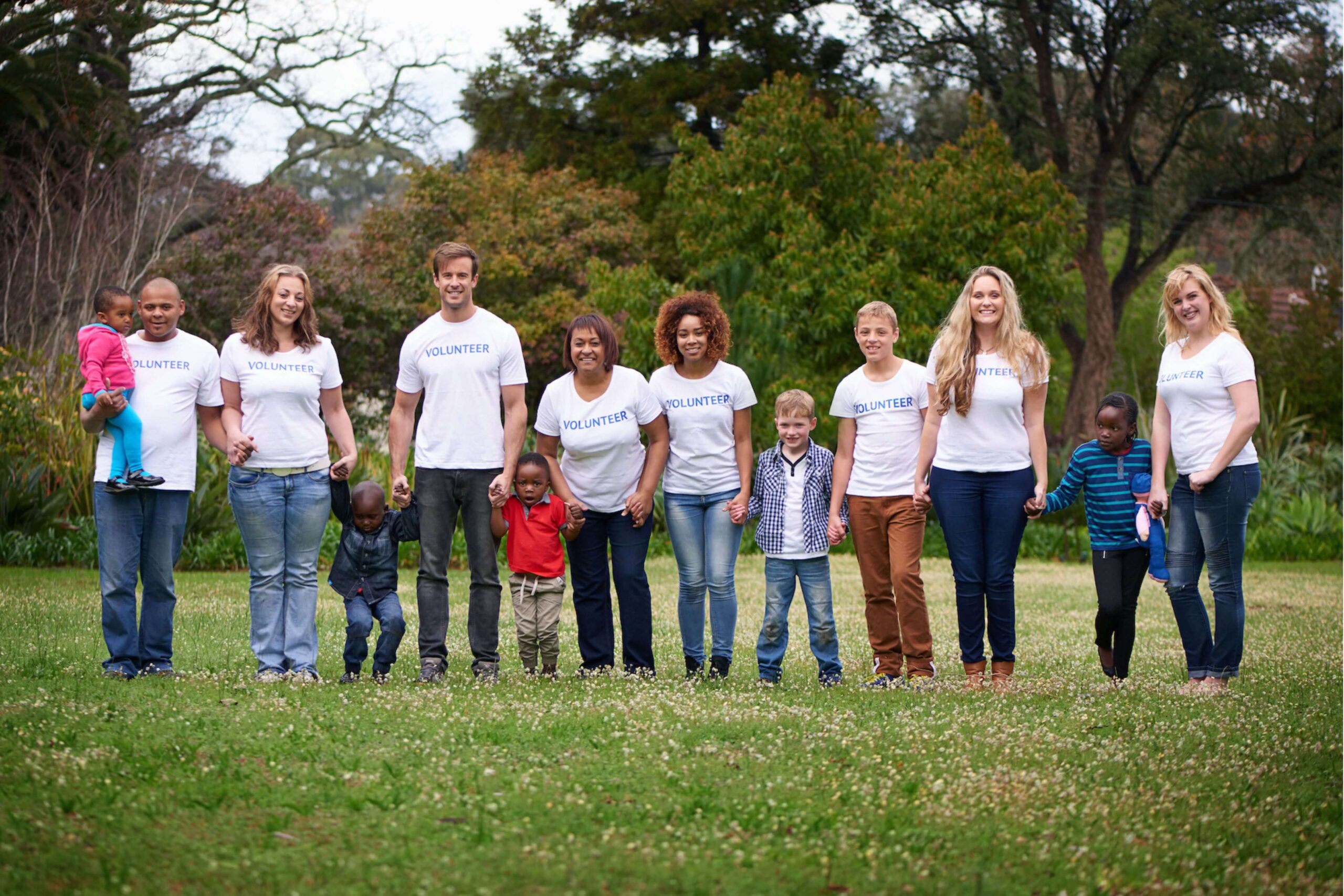 Group of volunteers and children standing in a park, holding hands, wearing white "Volunteer" shirts. Background features trees and greenery.