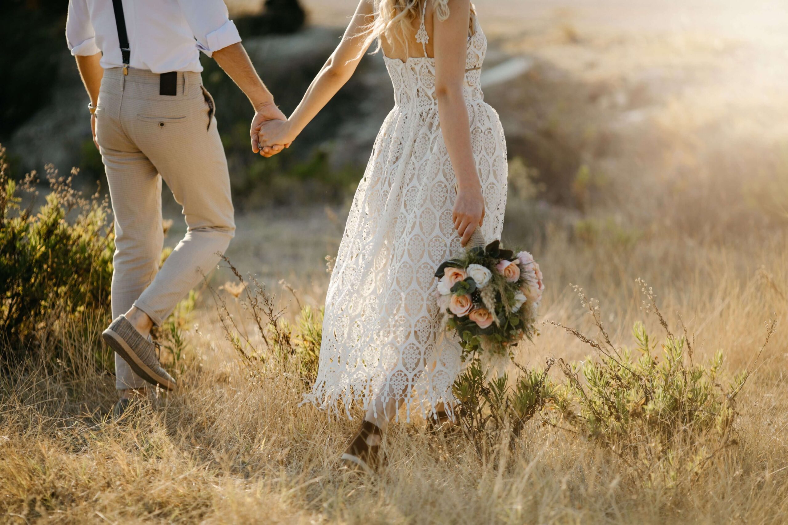 Two people holding hands, walking through a sunlit field. One person holds a bouquet of pink and white flowers, wearing a white lace dress.