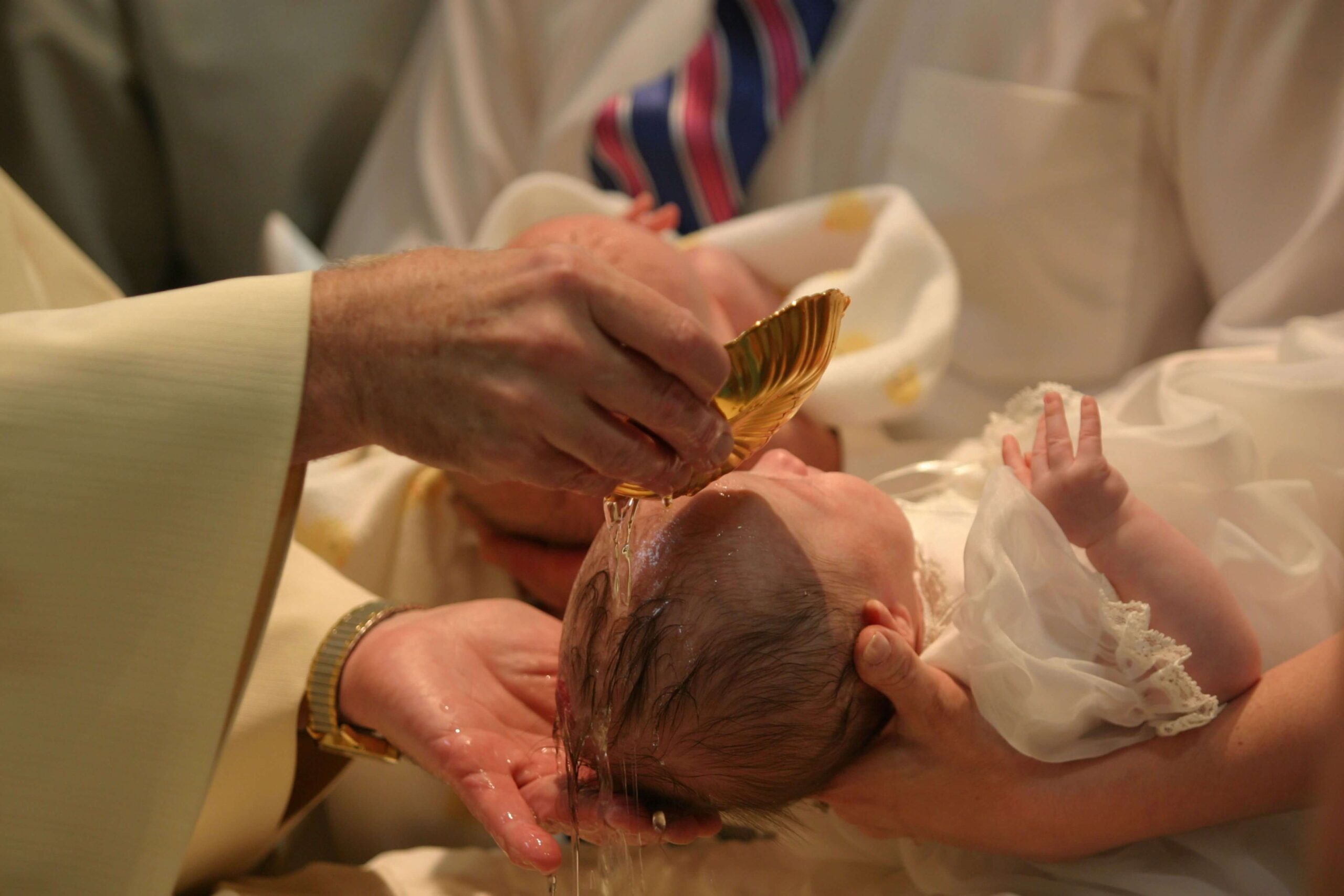 A person performs a baptism, pouring water from a golden shell over a baby's head, dressed in white, during a religious ceremony.