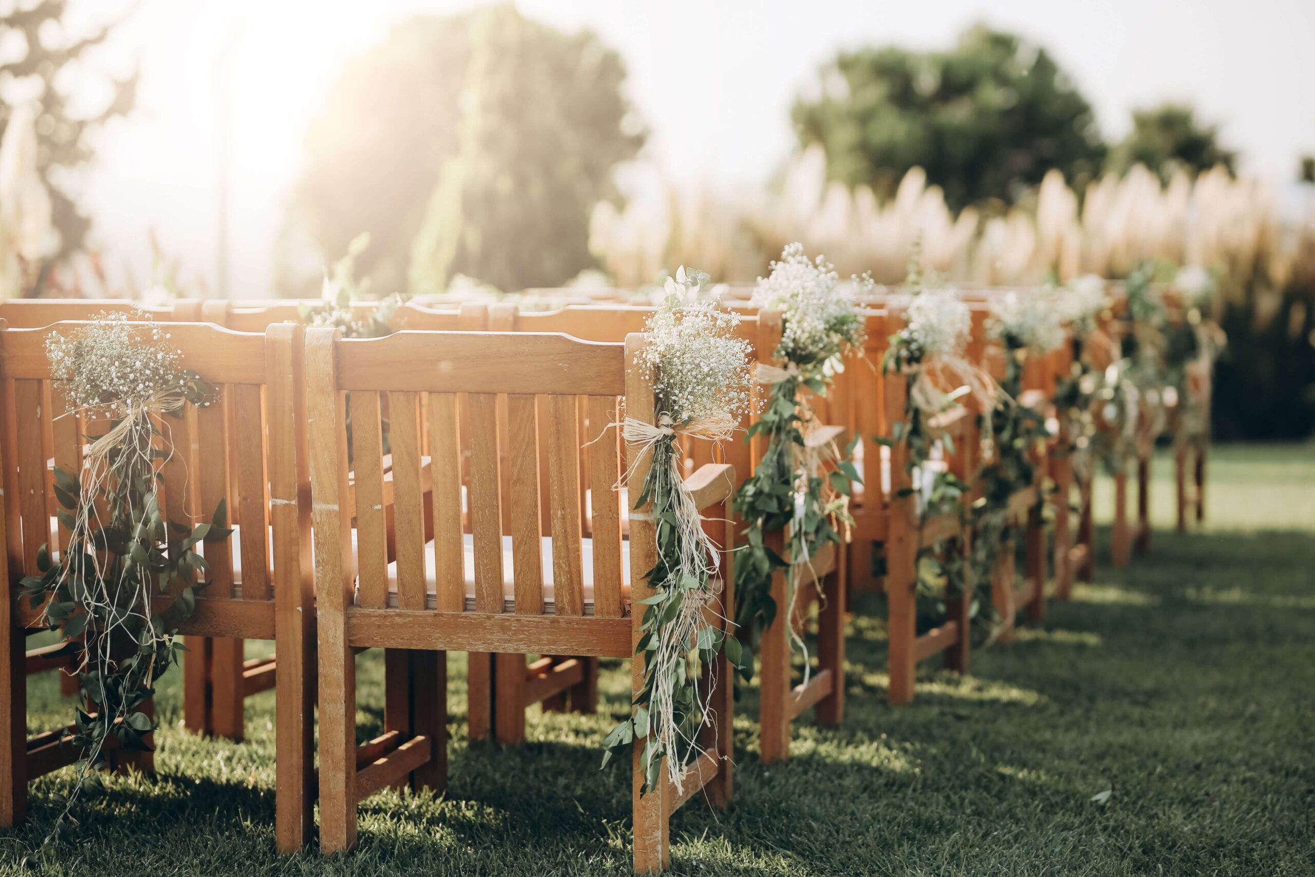 Sunlit outdoor wedding setup with rows of wooden chairs adorned with delicate flowers and greenery, creating an elegant and serene atmosphere.